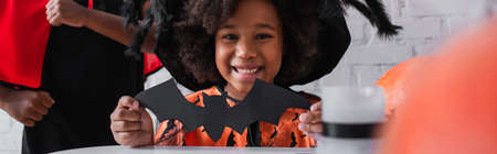 happy african american girl in halloween costume holding paper cut bat near brother, bannerの写真素材