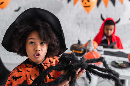 spooky african american girl in witch costume holding toy spider near blurred brotherの写真素材