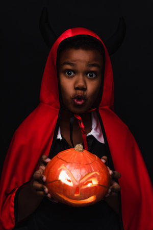 spooky african american boy in devil halloween costume holding carved pumpkin isolated on blackの写真素材