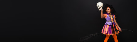 excited african american girl in witch halloween costume on top of broom holding skull isolated on black, bannerの写真素材