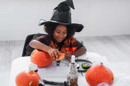 cheerful african american girl in witch halloween costume near pumpkins and cookies on tableの写真素材