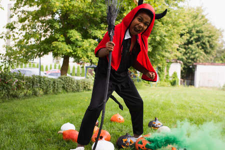 scary african american kid in devil halloween costume holding broom near pumpkins on lawn