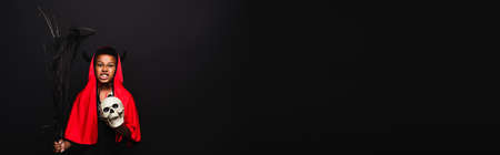 irritated african american boy holding broom and skull while grinning isolated on black, bannerの写真素材