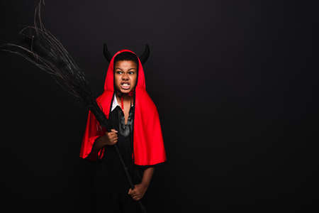 irritated african american boy holding broom and grinning isolated on black