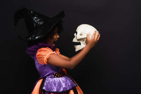 side view of excited african american girl in witch halloween costume holding skull isolated on blackの写真素材