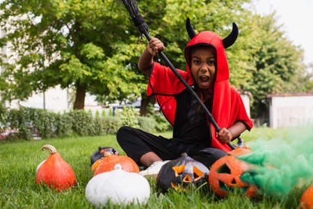 scary african american kid in devil halloween costume screaming while holding broom near pumpkins and sitting on lawnの写真素材
