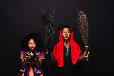 african american girl in halloween costume holding carved pumpkins near brother with brooms isolated on blackの写真素材
