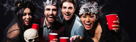 excited multiethnic friends with plastic cups laughing at camera during halloween party on black, bannerの写真素材