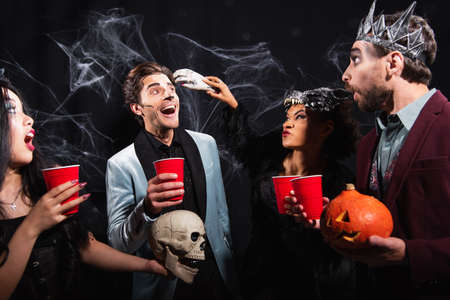 african american woman in wolf mask scaring man with toy hand near multiethnic friends on blackの写真素材