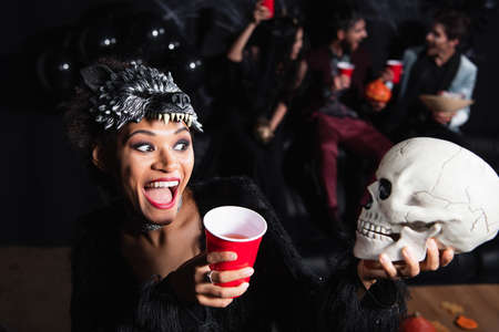 amazed african american woman in wolf mask looking at scary skull near blurred friends on black backgroundの写真素材