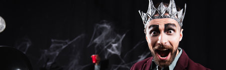 amazed man in vampire king halloween crown looking at camera on black, bannerの写真素材
