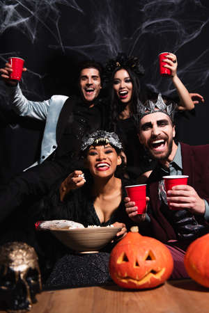 cheerful multiethnic friends in halloween costumes looking at camera while eating popcorn and drinking beer on blackの写真素材