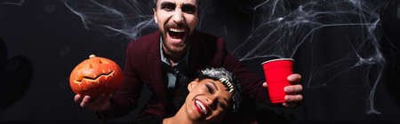 laughing man holding carved pumpkin and plastic cup near smiling african american woman in wolf mask, bannerの写真素材