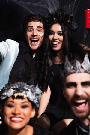 happy interracial friends in halloween costumes looking at camera on black, blurred foreground