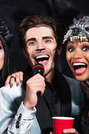 man in halloween makeup singing karaoke near excited african american woman in wolf mask on black