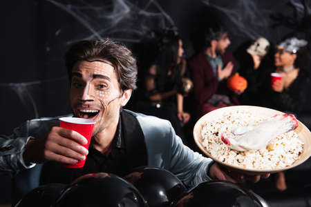 happy man in halloween makeup drinking beer near balloons and blurred friends on black backgroundの写真素材