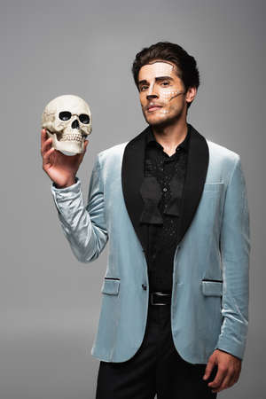 elegant man in elegant blazer and halloween makeup holding spooky skull isolated on greyの写真素材