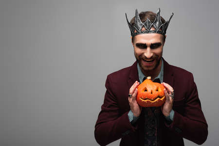 sarcastic man in vampire king halloween crown smiling and showing spooky pumpkin isolated on greyの写真素材