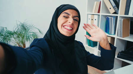 Smiling muslim businesswoman waving hand while taking selfie
