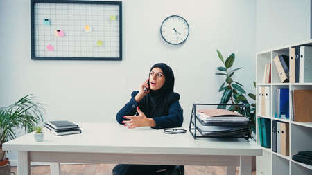Muslim businesswoman in hijab sitting at desk and talking on smartphone in officeの写真素材