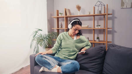 Smiling woman in wireless headphones using smartphone on couchの写真素材