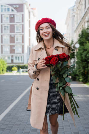 cheerful young woman in trench coat and red beret holding bouquet of roses on urban street of europeの写真素材