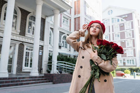 young woman in trench coat holding red roses and adjusting beret on urban street of europeの写真素材
