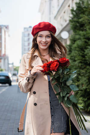 happy young woman in trench coat and red beret holding bouquet of roses on urban street of europeの写真素材