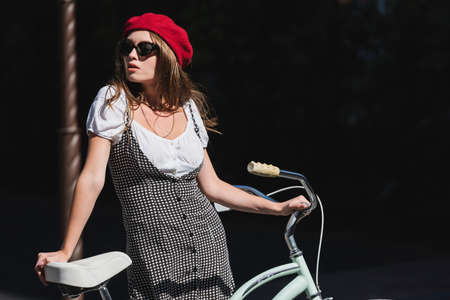 pretty young woman in sunglasses, beret and dress standing with bicycleの写真素材