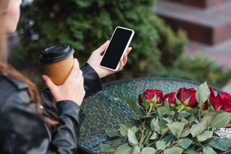 cropped view of young woman holding smartphone with blank screen and paper cup near roses on terrace of cafeの写真素材