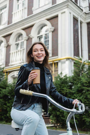 cheerful woman holding paper cup while riding bicycle on urban street of europeの写真素材