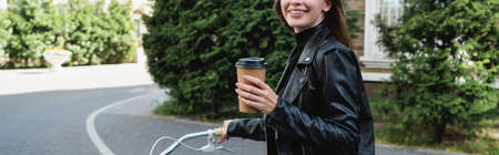 cropped view of happy woman holding paper cup while riding bicycle on urban street of europe, banner