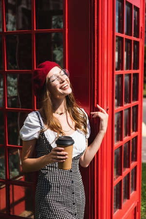 joyful woman in beret and eyeglasses holding paper cup near red phone boothの写真素材