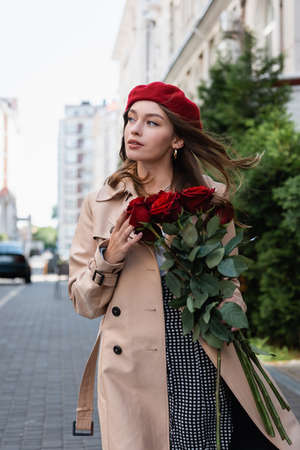pretty woman in trench coat and red beret holding bouquet of roses on urban street of europeの写真素材