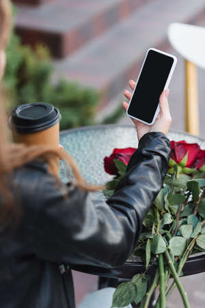 partial view of young woman holding smartphone with blank screen and paper cup on terrace of cafe
