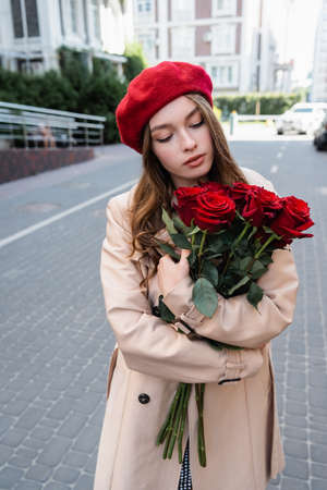 pretty woman in trench coat and beret looking at bouquet of red roses on urban street of europeの写真素材
