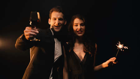 cheerful man holding glass of red wine and woman with sparkler looking at camera on blackの写真素材