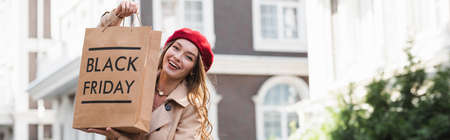 cheerful young woman in red beret and trench coat holding shopping bag with black friday lettering outside, bannerの写真素材