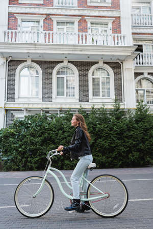 full length of joyful and stylish woman riding bicycle on urban street of europeの写真素材