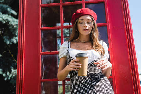 pretty woman in beret and eyeglasses holding paper cup near red phone boothの写真素材