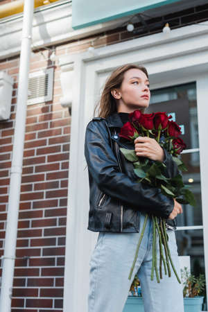 young stylish woman in leather jacket holding red roses on urban street in europeの写真素材