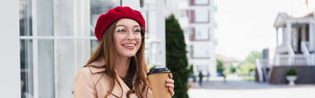 happy young woman in red beret and beige trench coat holding paper cup, bannerの写真素材