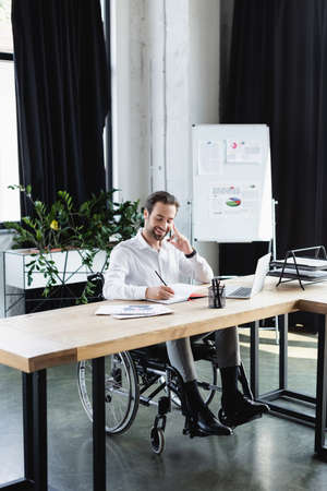 positive disabled businessman writing in notebook during conversation on smartphone in office