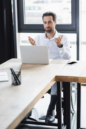 young businessman pointing with hands at laptop during video conference in office