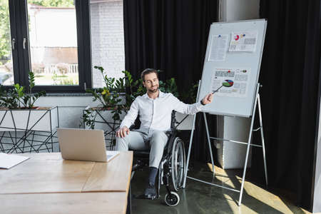 disabled businessman in wheelchair pointing at flip chart during video call on laptop in officeの写真素材