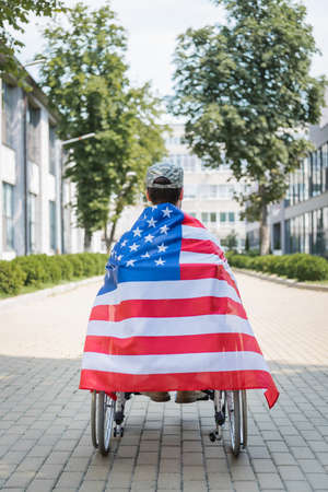 back view of veteran in wheelchair, wrapped in usa flag on city alleyの写真素材