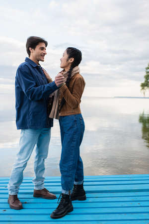 Side view of interracial couple holding hands on pier near lake