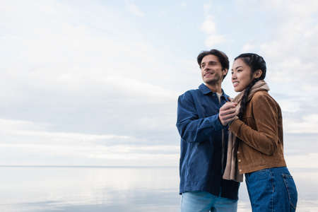 Smiling multiethnic couple holding hands near lake and sky at background