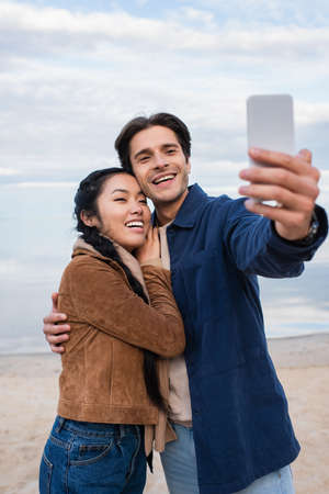 Smiling interracial couple hugging and taking selfie on blurred smartphone on beach