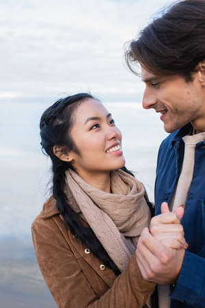 Smiling multiethnic couple holding hands while looking at each other near lakeの写真素材